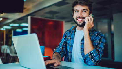 Fototapeta na wymiar Portrait of happy Caucasian hipster guy smiling at camera while using cellular for positive calling in coworking space, cheerful male IT professional with laptop for programming posing while phoning