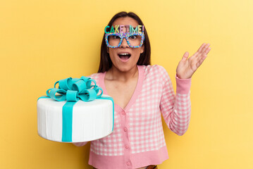 Young mixed race woman holding a cake isolated on yellow background receiving a pleasant surprise, excited and raising hands.