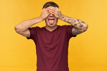 Naklejka premium Indoor shot of funny young male, closed his eyes with arms, shows tongue while waiting for surprise, isolated over yellow background