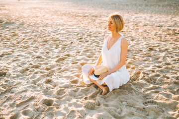 Fototapeta na wymiar Blonde woman wearing long white dress sit at empty beach with her eyes closed