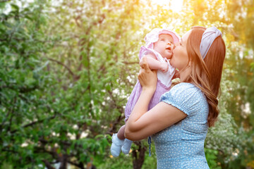 Obraz premium Fair haired young woman in blue dress holds in arms little daughter and kisses baby cheek with happy smile in green park on spring day, sunlight