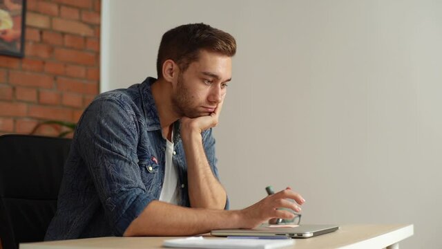 Side view of bored young businessman tapping on cover of laptop with fingers sitting at desk in office. Nervous sad upset male tapping waiting for client in conference room. Shooting in slow motion.