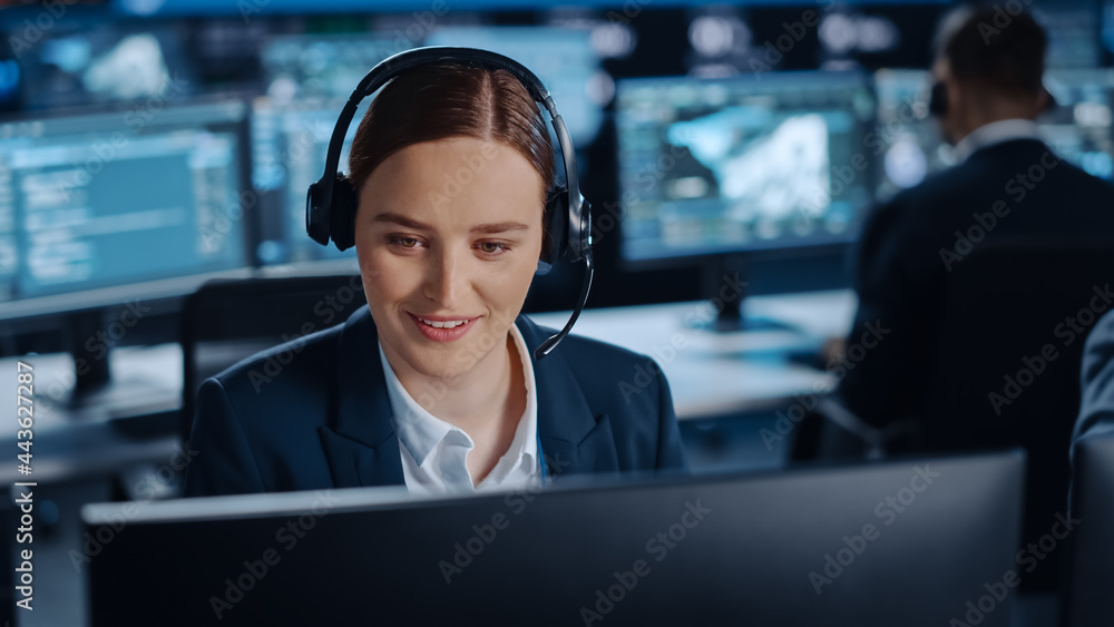 Poster Happy Beautiful Technical Customer Support Specialist is Talking on a Headset while Working on a Computer in a Dark Monitoring and Control Room Filled with Colleagues and Display Screens.