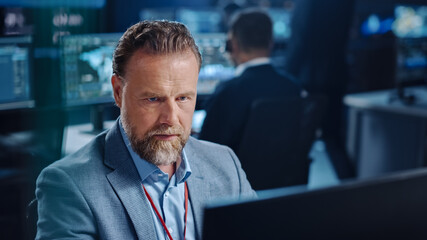 Portrait of Serious Professional Technical Controller Sitting at His Desk with Multiple Computer Displays Before Him. In the Background His Colleagues Working in System Control and Monitoring Center. © Gorodenkoff