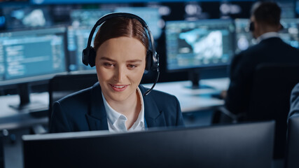 Happy Beautiful Technical Customer Support Specialist is Talking on a Headset while Working on a Computer in a Dark Monitoring and Control Room Filled with Colleagues and Display Screens.