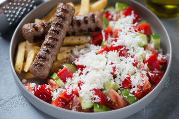 Close-up of serbian shopska salad with grilled cevapi and fries in a grey plate, selective focus