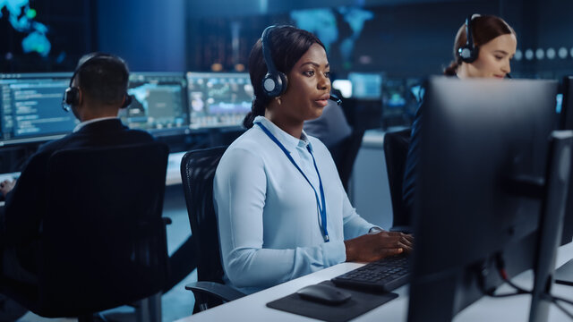 Happy Beautiful Technical Customer Support Specialist is Talking on a Headset while Working on a Computer in a Call Center Control Room Filled with Colleagues, Display Screens and Data Servers.