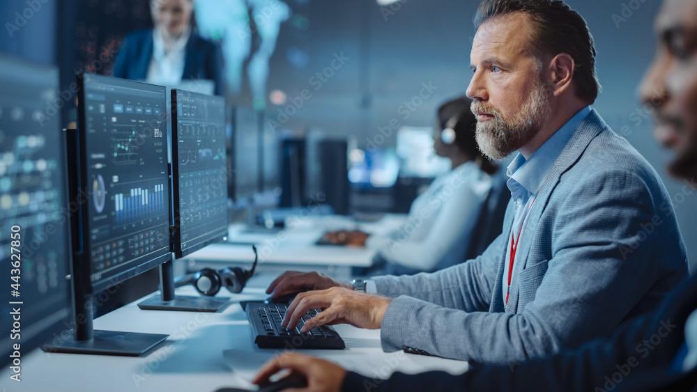 Poster Portrait of Serious Professional Technical Controller Sitting at His Desk with Multiple Computer Displays Before Him. In the Background His Colleagues Working in System Control and Monitoring Center.