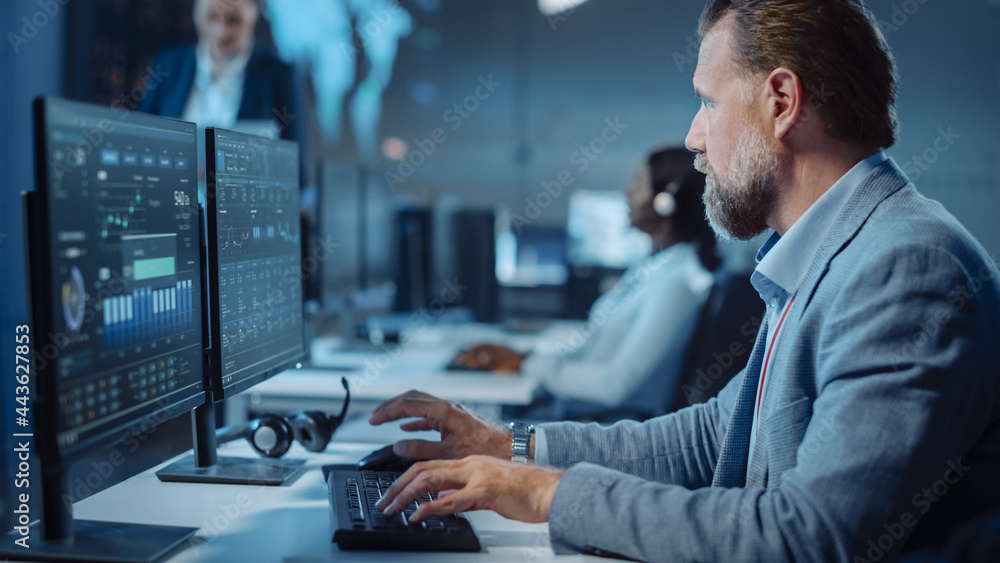 Poster Portrait of Serious Professional Technical Controller Sitting at His Desk with Multiple Computer Displays Before Him. In the Background His Colleagues Working in System Control and Monitoring Center.