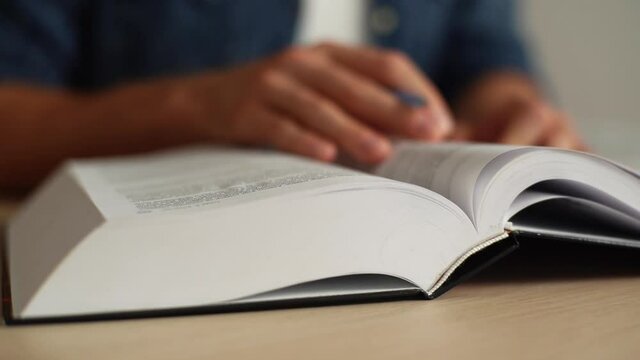 Close-up hands of unrecognizable man turning pages of reference book, slow motion, blurred background. Male fingers over page of book, looking for right information of big encyclopedia.
