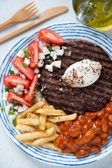 Blue and white plate with bbq balkan pljeskavica, kaymak and bryndza cheese, red tomatoes, fries and prebranac, vertical shot, top view