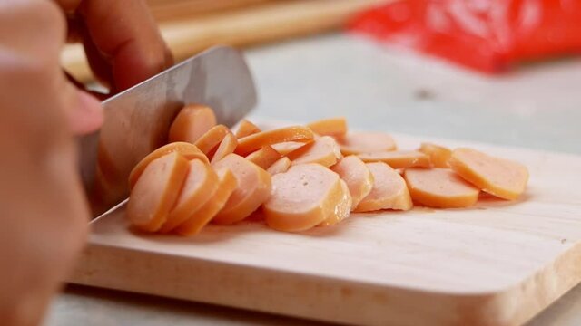 Close up of a knife cuts the sausage into small pieces. Mother holds her daughter's hand to teach her how to cut sausages to prepare them for making Borona sauce over spaghetti at home.