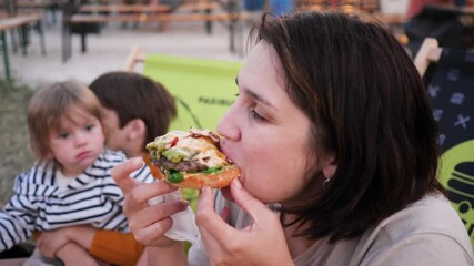 Woman eating a tasty juicy part of a meat burger while her children watching her at street food trucks festival