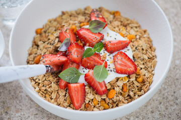 Granola served with fresh strawberry and yogurt in a white bowl, closeup on a beige marble background, selective focus