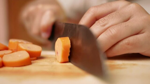 Close up of a knife cuts the sausage into small pieces. Mother holds her daughter's hand to teach her how to cut sausages to prepare them for making Borona sauce over spaghetti at home.