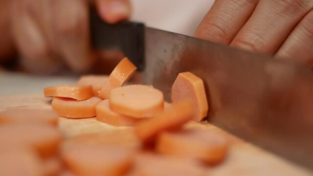 Close up of a knife cuts the sausage into small pieces. Mother holds her daughter's hand to teach her how to cut sausages to prepare them for making Borona sauce over spaghetti at home.
