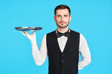 Attractive young waiter holding empty silver tray over blue background - obrazy, fototapety, plakaty