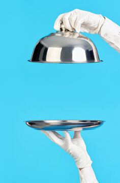 Elegant waiter's hands in white gloves holding silver tray and cloche on blue background