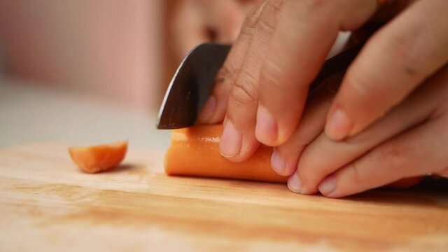 Close up of a knife cuts the sausage into small pieces. Mother holds her daughter's hand to teach her how to cut sausages to prepare them for making Borona sauce over spaghetti at home.