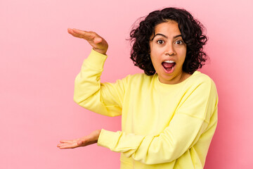 Young curly latin woman isolated on pink background shocked and amazed holding a copy space between hands.