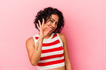 Young curly latin woman isolated on pink background winks an eye and holds an okay gesture with hand.