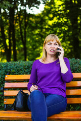 Defocus wonder caucasian blond woman talking, speaking on the phone outside, outdoor. 40s years old woman in purple blouse in park. Adult person using phone. Vertical. Out of focus