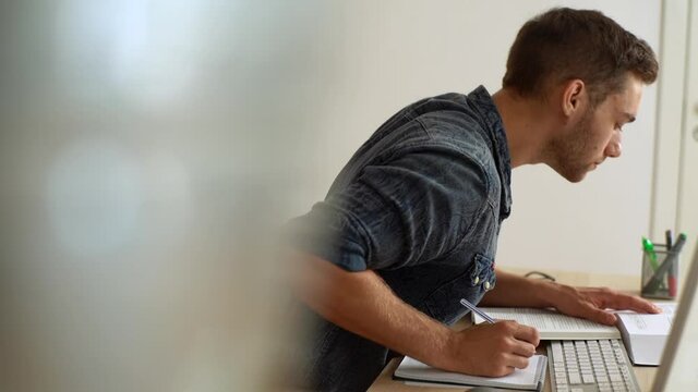 Side view of university male student in casual clothes studying taking notes from book while sitting at desk. Tracking shot of focused Caucasian guy writing notes during homework, slow motion.