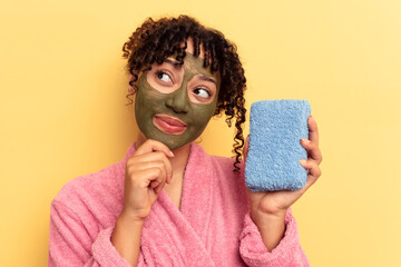 Young mixed race woman wearing a pink bathrobe holding a shower sponge isolated on yellow background looking sideways with doubtful and skeptical expression.