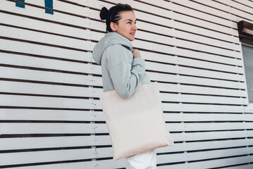 Pretty woman in hoodie with white cotton bag on her shoulder. Mockup and zero waste concept.