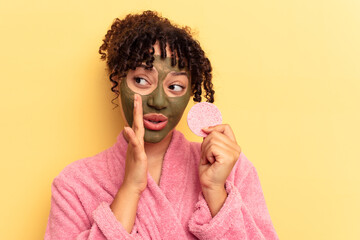 Young mixed race woman wearing a bathrobe holding a make-up remover sponge isolated on yellow background is saying a secret hot braking news and looking aside