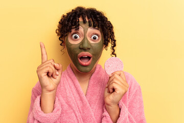 Young mixed race woman wearing a bathrobe holding a make-up remover sponge isolated on yellow background having an idea, inspiration concept.