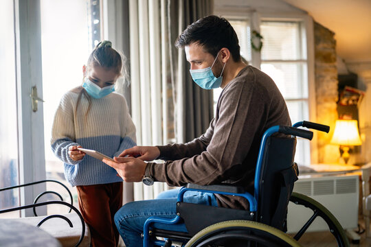 Father with disability in wheelchair using tablet at home with child while wearing masks.