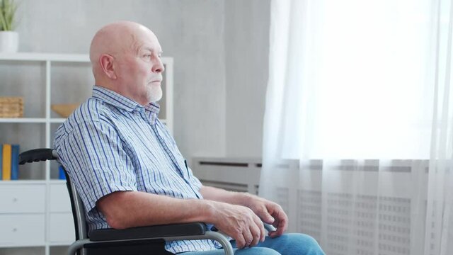 Disabled old man is sitting in a wheelchair at home alone. A handicapped person in a wheelchair is looking out the window. Paralysis and disability.