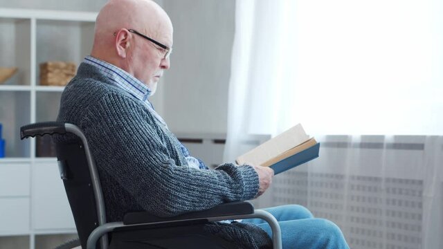 Disabled old man is sitting in a wheelchair at home alone. A handicapped person in a wheelchair is reading the book. Paralysis and disability.