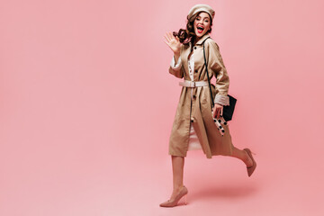 Portrait of girl in midi trench coat and hat. Woman holds her bag, waves hand and runs on pink background