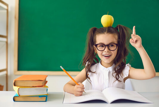 Back to school concept. Little girl schoolgirl with glasses raised her finger up has an idea on the background of a greenboard in elementary school. Education learning lessons for children.