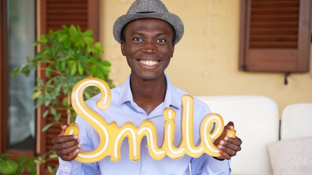 Portrait of a cheerful optimistic modern African-American man with a trendy hat smiles and shows a label with the word smile. 