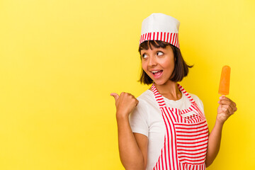 Young mixed race ice cream maker woman holding an ice cream isolated on yellow background points with thumb finger away, laughing and carefree.