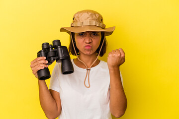 Young mixed race explorer woman holding binoculars isolated on yellow background showing fist to camera, aggressive facial expression.