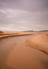 Dunstanburgh Castle in Northumberland, England, taken with the river meandering in the foreground & with cloudy skies overhead