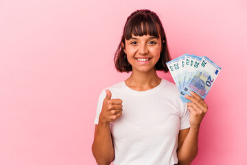 Young mixed race woman holding bills isolated on pink background smiling and raising thumb up