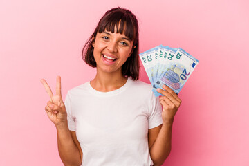 Young mixed race woman holding bills isolated on pink background joyful and carefree showing a peace symbol with fingers.