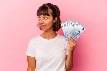 Young mixed race woman holding bills isolated on pink background looks aside smiling, cheerful and pleasant.