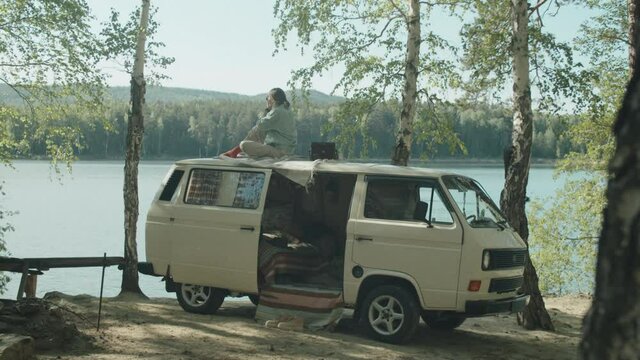 Wide shot of woman sitting on roof of camper van and taking pictures of lake and her dog with vintage camera