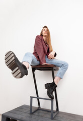 Fashionable vogue woman model dressed in jeans, leather jacket and corset sitting on chair, white background