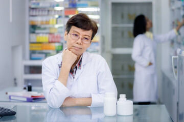 female pharmacist standing back couunter with hand on chin in pharmacy