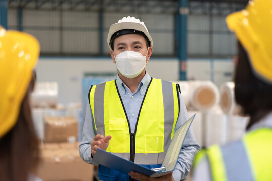 Asian Male Supervisor Engineer In Wearing Helmet Safety And Hygienic Mask To Protect Coronavirus Stands With Documents Explaining Action Plan To Teamwork At Warehouse Factory Industrial.