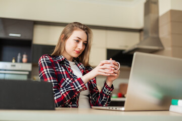 Young attractive woman looking at the laptop screen while sitting at the table and drinking coffee in the living room