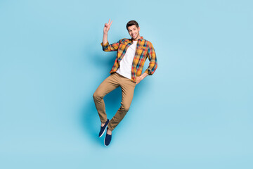 Photo of funky attractive young gentleman wear checkered shirt jumping high showing v-sign smiling isolated blue color background
