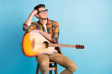 Photo of funny dreamy young gentleman wear checkered shirt holding guitar looking empty space smiling isolated blue color background